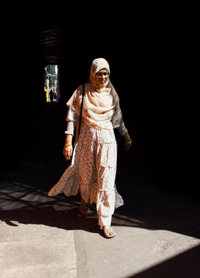 An Indian woman in traditional clothing stepping into a section of bright sunlight from dark shadow, with a distant window of light behind her framing someone's silhouette.